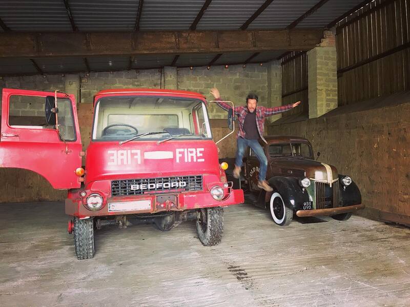 Man jumping off an old Bedford fire truck with a vintage Ford truck in the background