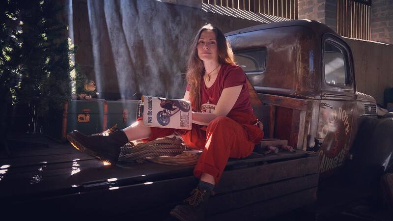 Woman in overalls reading a car magazine whilst sitting on the back of an old pick up truck inside a workshop