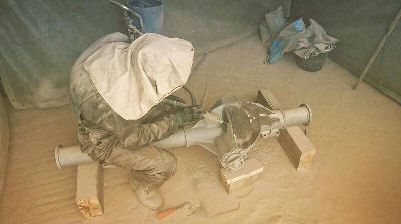 Man in tent with hood on sandblasting the axle of a Bedford truck