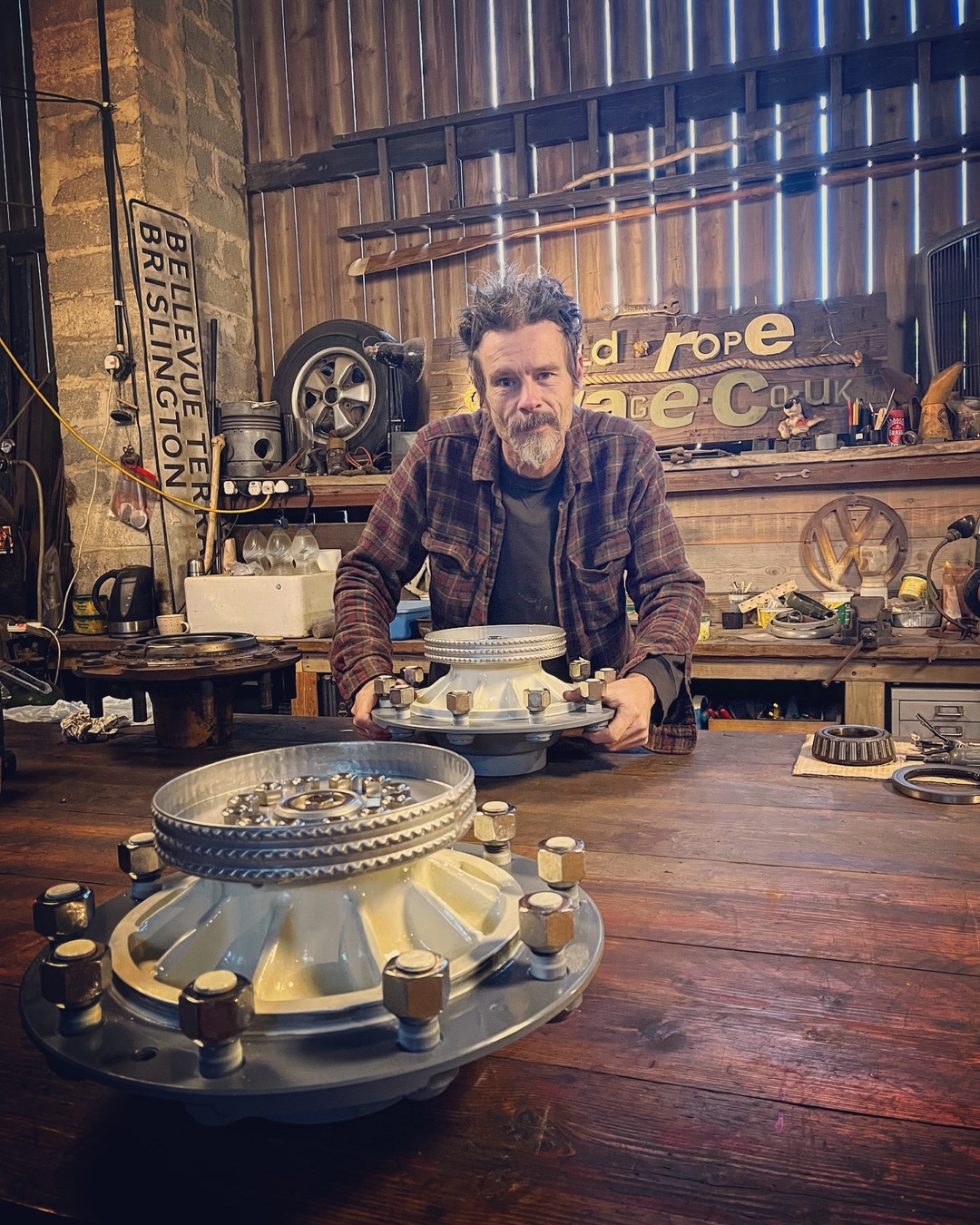 Man standing behind a workbench with 2 partially restored and painted Bedford truck wheel hubs