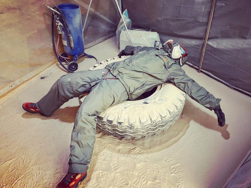Woman in protective clothing lying on a truck tyre with sandblasting equipment