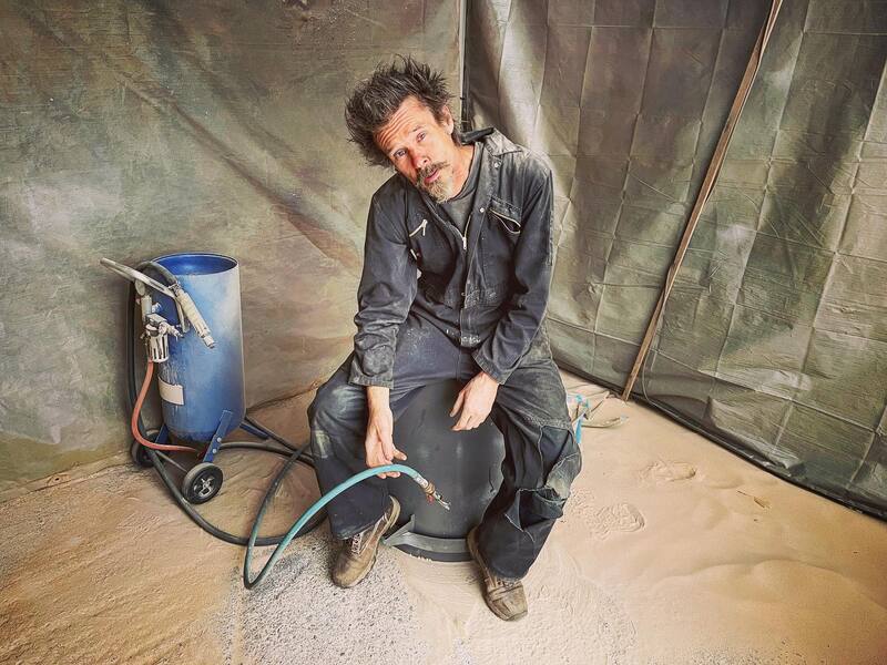 Man in overalls sitting on an upside down bin covered in sand with a sandblaster in his hand