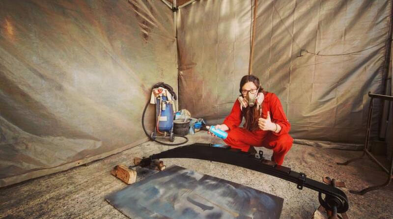 Woman in orange overalls and face mask in a spray tent spray painting leaf springs
