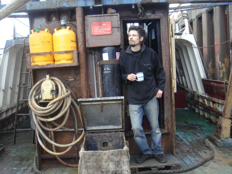 Man with a mug of tea in hand standing on deck outside the door to the wheelhouse of an old wooden fishing trawler