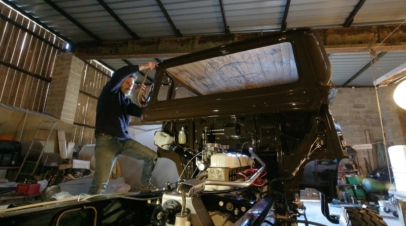 Man standing on the back of a brown classic Bedford truck, fitting a grab handle to the cab