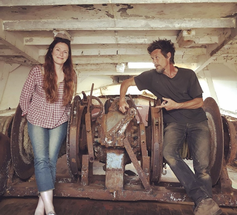 Man and woman stand in front of a large boat winch inside a wooden boat