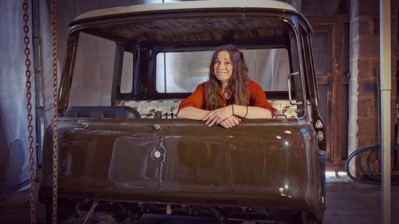 Woman in orange overalls sits inside a brown Bedford truck cab which is being restored