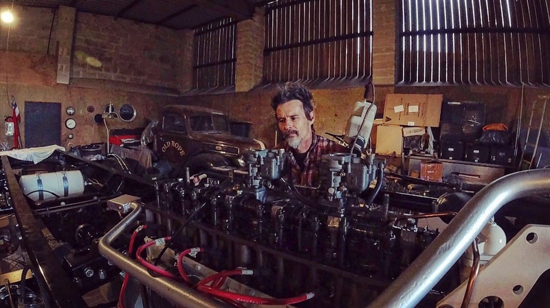 Man standing over a Bedford truck petrol engine with the rocker cover removed