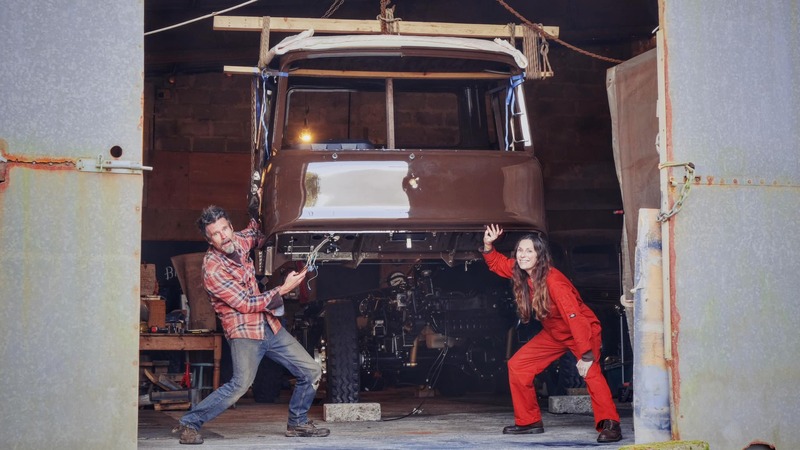 Man and woman under a Bedford truck cab which has been winched in to the air