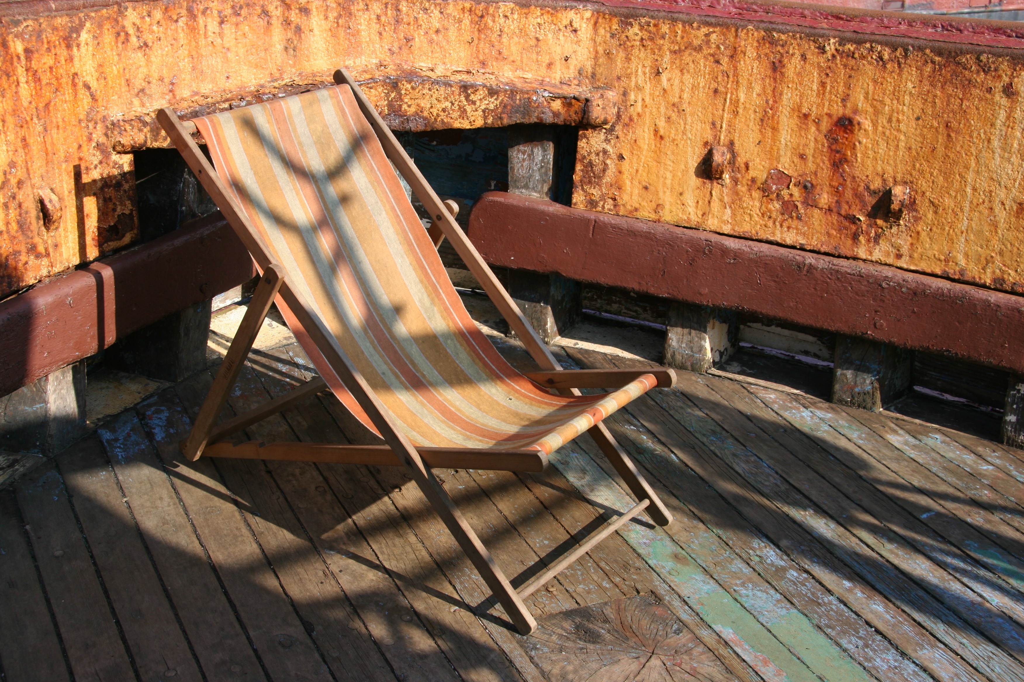 An old 1970s style wooden deckchair on a wooden boat deck with a rusty metal wall in the background