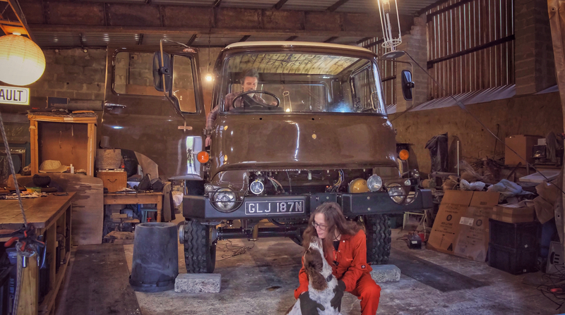 Brown Bedford truck in a workshop with a man sitting in the driving seat and a woman playing with a dog 