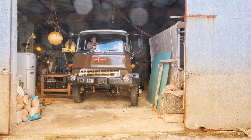 Man driving a brown restored Bedford truck out of a workshop